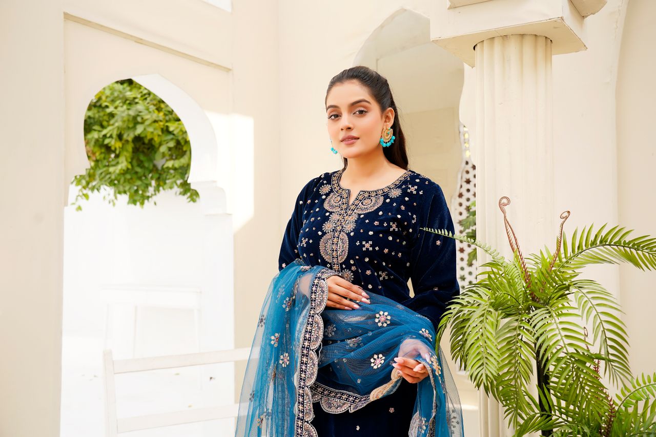 A woman poses elegantly in a blue embellished traditional dress indoors beneath classic architecture.